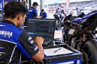 Mekanik balap motor menganalisis data ECU menggunakan laptop untuk mengevaluasi performa mesin di paddock. Salin alt text