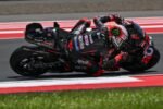 Ilustrasi AI pembalap Aprilia MotoGP dengan motor hitam merah saat menikung ekstrem di lintasan balap.