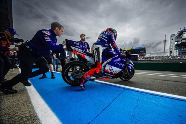 Ilustrasi AI pembalap MotoGP di atas motor balap biru bersama kru tim di area pit lane sirkuit.
