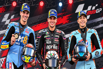 Ilustrasi AI tiga pembalap MotoGP mengenakan seragam balap dan topi Michelin sambil membawa helm di podium.